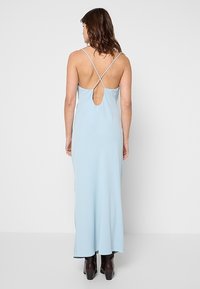 Femme portant une robe longue bleu clair dos nu avec de fines bretelles croisées et des bottes à talons noires, debout devant un fond blanc uni.