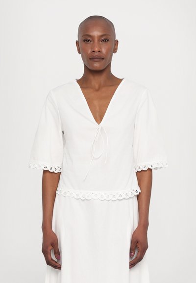 Blouse blanche avec un décolleté en V profond, des manches courtes et de la dentelle le long de l'ourlet et des manches. Fabriquée en tissu léger avec un détail noué.