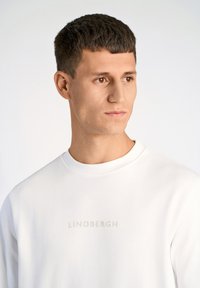 Vit sweatshirt med präglad "LINDBERGH"-text på bröstet, rund hals och en slät, mjuk textur. Modellen har kort hår.