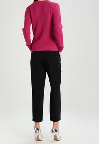 Frau in einem fuchsiafarbenen, gerippten Pullover, schwarzen knöchellangen Hosen und silbernen High Heels steht mit den Händen in den Taschen, mit dem Rücken zur Zuschauerin.