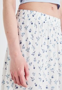 Hvit skjørt med teksturert stoff, med et blått blomsterdesign. Elastisk midje er synlig, med en hånd som hviler på stoffet.