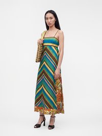 Femme portant une robe longue colorée rayée et fleurie avec bretelles spaghetti, tenant un sac tissé et des sandales noires à talons hauts.