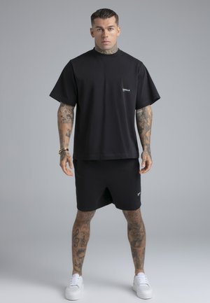 Bărbat cu tatuaje, purtând un tricou negru supradimensionat și pantaloni scurți asortați, stând cu brațele relaxate pe lângă corp, pantofi sport albi, fundal gri.