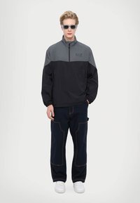 Sort og grå vindjakke med halvhøj lynlås foran, elastiske manchetter og logo på brystet, parret med mørkeblå cargo-bukser og hvide sneakers.