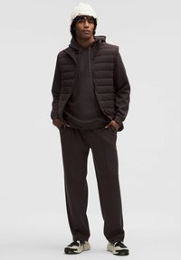 Dunkelbraune gepolsterte Weste über einem passenden Hoodie und einer Jogginghose, kombiniert mit schwarz-weißen Sneakers. Weicher Stoff, lässige Passform.