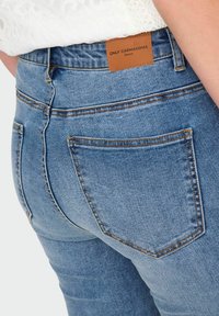 Mėlyna džinsinė kelnė su aukštu juosmeniu, matomu siūliniu, prekės ženklo odiniu etiketė ant nugaros ir dviem užpakalinėmis kišenėmis.