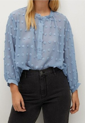 Frau trägt eine hellblaue, strukturierte Bluse mit kleinen erhabenen Punkten, am Kragen geknöpft, in eine hoch taillierte schwarze Jeans gesteckt.