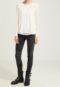 Bílá blůza s dlouhým rukávem a texturovaným živůtkem, v kombinaci s těsnými černými denimovými kalhotami a černými kotníkovými botami s několika sponami.