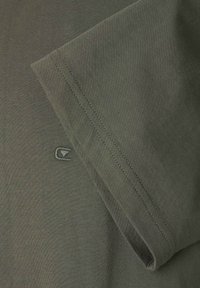 Dettaglio ravvicinato di una maglietta a maniche corte di colore verde oliva con orlo doppio cucito e piccolo logo ricamato vicino al bordo della manica.