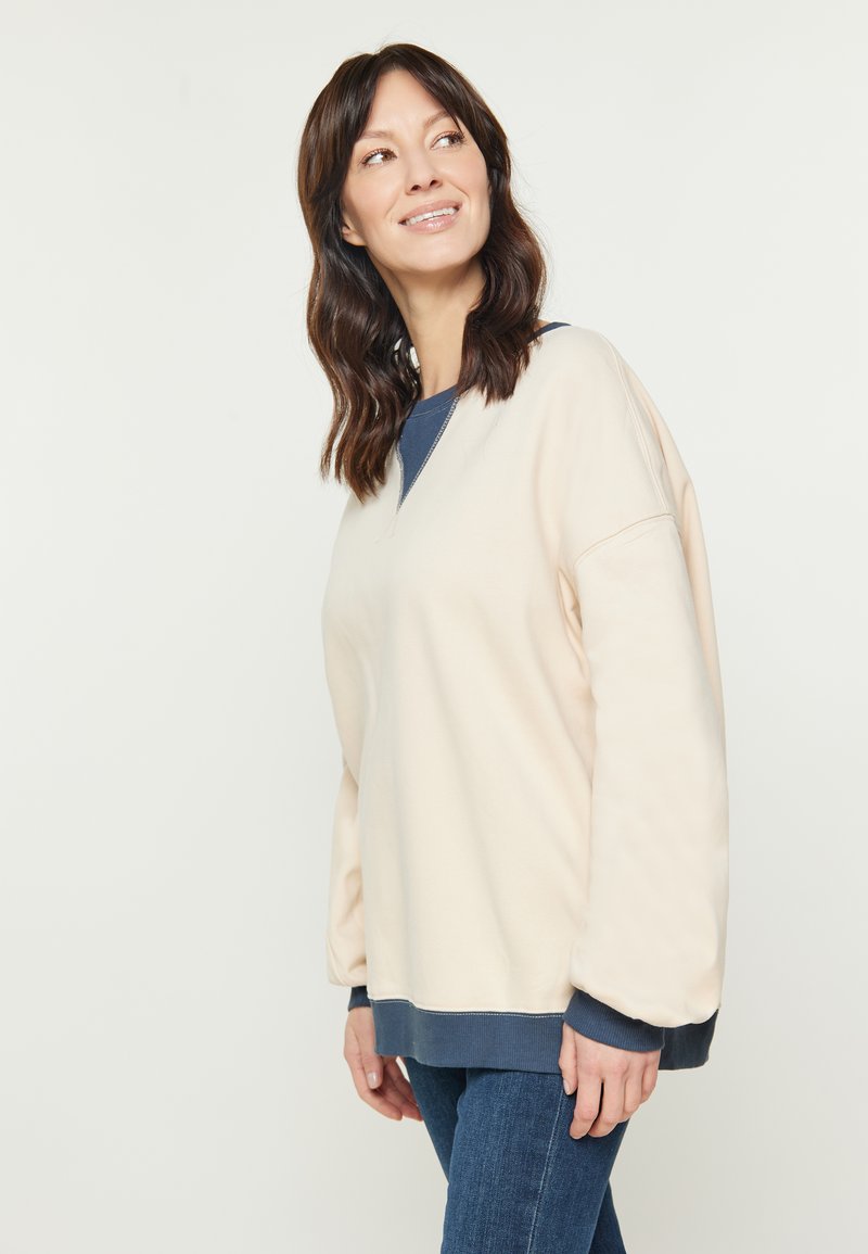 Felpa beige con scollo a V e maniche lunghe e oversize, con un bordo inferiore blu scuro e polsini. Texture liscia.