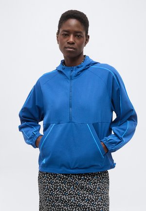 Persoon draagt een blauwe capuchontrui met zakken aan de voorkant en een zwarte rok met witte en blauwe patronen, staand tegen een effen achtergrond.