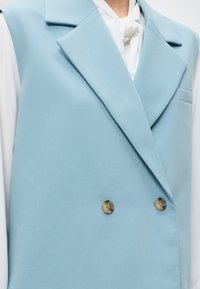 Gilet bleu clair croisé à larges revers porté sur un chemisier blanc avec un detail noué au col, avec deux boutons marron.