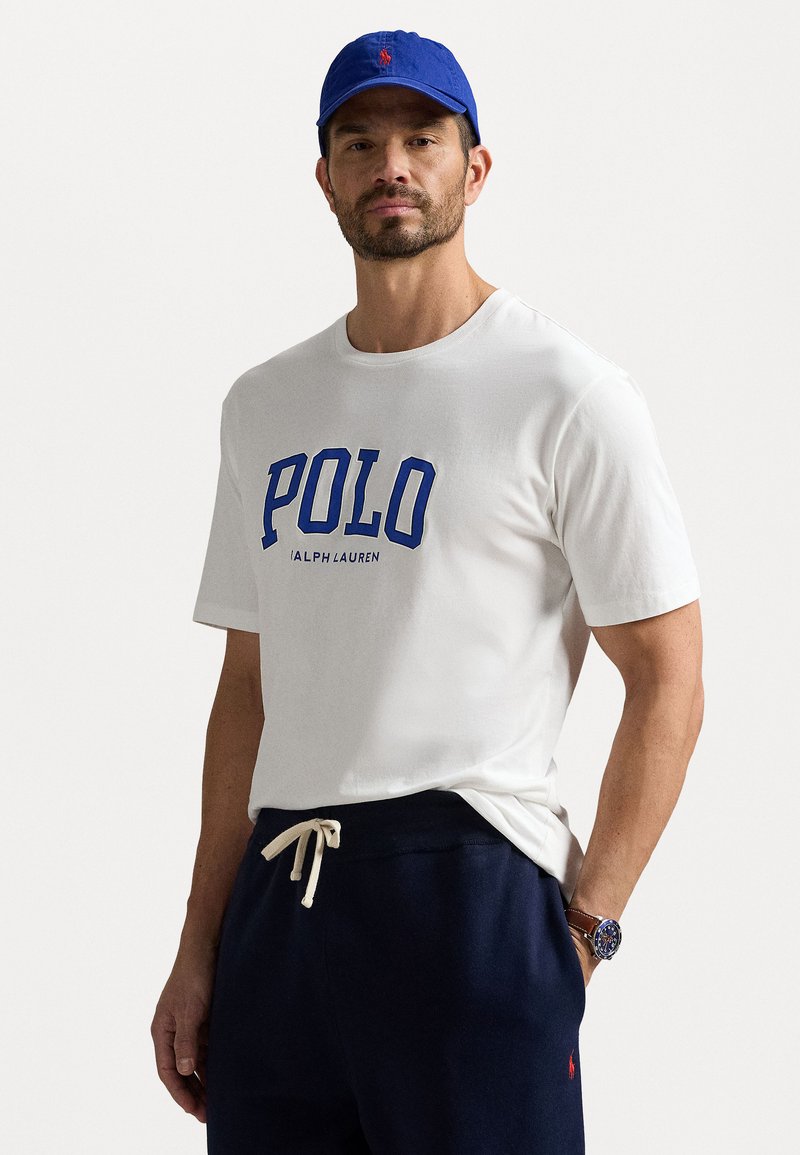 T-shirt en coton blanc avec logo "POLO" bleu et texte "RALPH LAUREN". Associé à un pantalon de survêtement marine et une casquette bleue, avec un détail de logo.