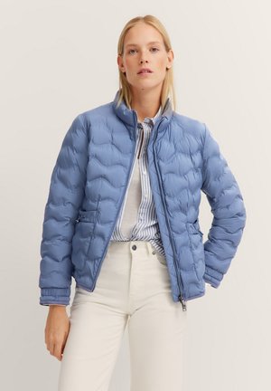 Helle blaue Pufferjacke mit gestepptem Design, Reißverschluss, hohem Kragen und seitlichen Taschen, getragen über einem gestreiften Hemd und hellen Hosen.