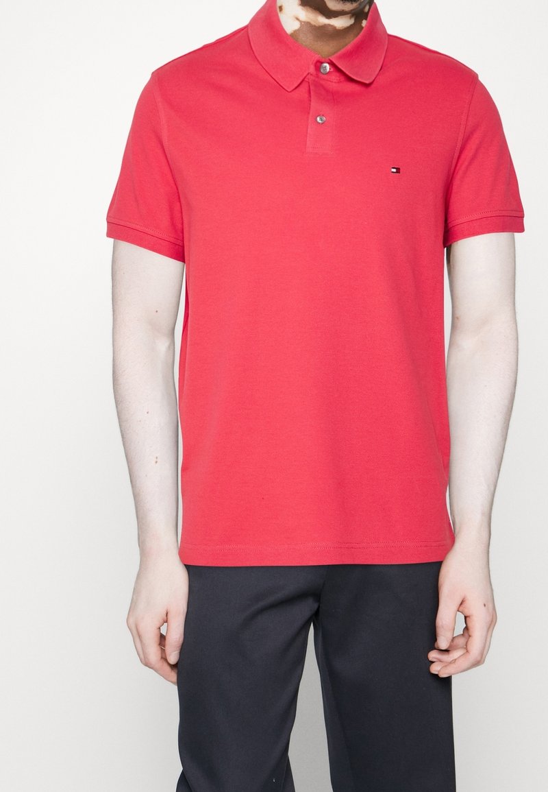Polo en coton rouge avec un col classique, des manches courtes et un petit logo sur la poitrine. Assorti à un pantalon foncé.