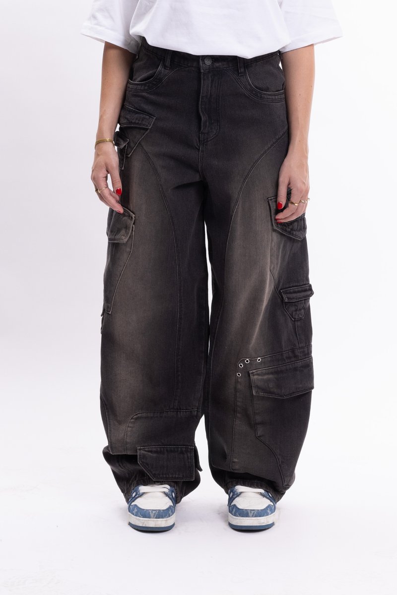 Pantalones cargo de mezclilla negra con múltiples bolsillos, corte holgado y detalles metálicos. Llevados con zapatillas de deporte de patrón azul y blanco.