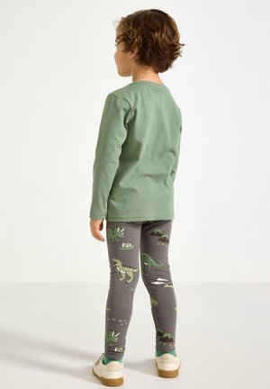 Barn med krøllet hår iført en grøn langærmet trøje og grå leggings med dinosaurprint, stående med ryggen mod på en ensfarvet baggrund.