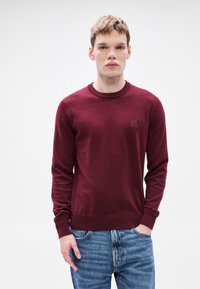 Πλεκτό πουλόβερ μπουρντό με στρογγυλή λαιμόκοψη, μακριά μανίκια και υφή υφάσματος. Διαθέτει μικρό λογότυπο στο στήθος.