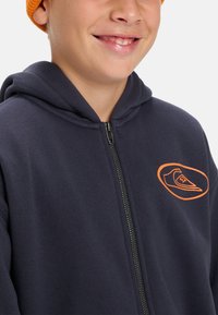 Navy zip-up hoodie made of soft fabric with a front pocket, featuring an orange shoe graphic on the left chest area.