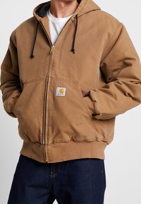 Brun zip-up hoodie i bomull, med dragsko i huvan, framfickor, ribbade muddar och en liten märkt patch på bröstet.