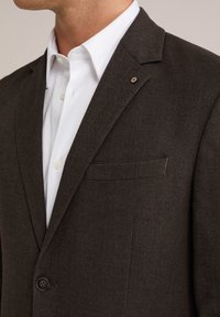 Detailný záber osoby v tmavo hnedom saku cez biele košeľové tričko, na ktorom sú vidieť klopy saka, vrecko a detaily gombíka.