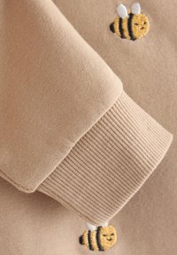 Tissu de sweat-shirt beige avec des poignets côtelés, orné d'abeilles brodées jaunes et noires. Texture douce et design décontracté.