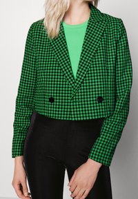 Lyhennetty bleiseri vihreässä houndstooth-kuviossa, jossa on notche-läppä, kaksoispainikkeet ja mustat yksityiskohdat, yhdistettynä tekstuurisiin mustiin korkeavyötäröisiin housuihin.