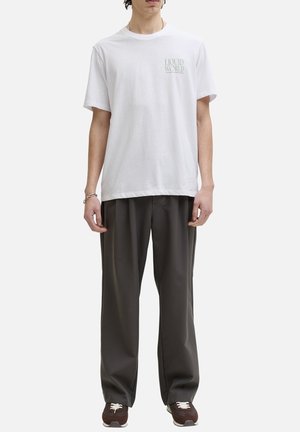Jeune homme portant un t-shirt blanc avec le texte « Liquid World » sur la poitrine, un pantalon ample gris foncé et des baskets marron, debout devant un fond uni.