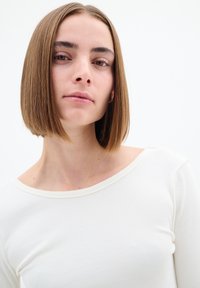 Bílé žebrované tričko s dlouhým rukávem a širokým výstřihem, s jemnou strukturou. Modelka má rovné středně hnědé vlasy stylizované do bobu.