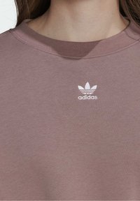 Dammig rosa sweatshirt i bomullsblandning. Har rund halsringning, ribbad nederkant och en vit Adidas-logotyp med tre ränder ovanför.