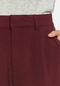 Burgundijas bikses ar gludu tekstūru, ar sānu kabatām un plakanu jostasviju. Redzamā šuvuma detaļas piešķir papildus akcentu.