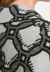 Tessuto in misto lana grigia con motivo geometrico nero. Il design presenta forme intrecciate e dettagli testurizzati sulla superficie.