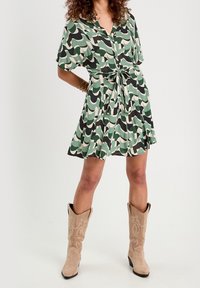 Robe verte et beige à motifs avec une taille nouée, des manches courtes et une jupe évasée, associée à des bottes de cow-boy beiges avec des coutures décoratives.