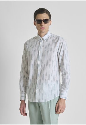 Hombre con gafas de sol oscuras, camisa blanca con rayas verticales grises y pantalones verde claro, de pie frente a un fondo liso.