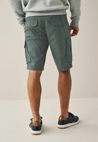 Shorts cargo verts avec poches latérales et rabats arrière, assortis à des chaussures bleues avec des accents bruns, le tout sur un fond neutre.