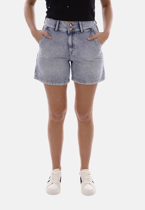 Persona che indossa pantaloncini di jeans azzurri, top nero, sneakers bianche, in piedi con le mani nelle tasche contro uno sfondo bianco.