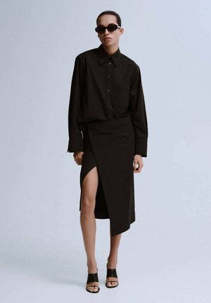 Czarna koszula na guziki i asymetryczna spódnica z wysokim rozcięciem na udo, zestawione z czarnymi sandałami z odkrytymi palcami oraz okularami przeciwsłonecznymi. Gładka, matowa faktura.