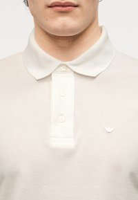 Άνδρας που φοράει λευκό πόλο μπλουζάκι με κουμπιά και μικρό κεντητό λογότυπο στο αριστερό στήθος, φαίνεται από τον λαιμό μέχρι το μέσο του κορμού.