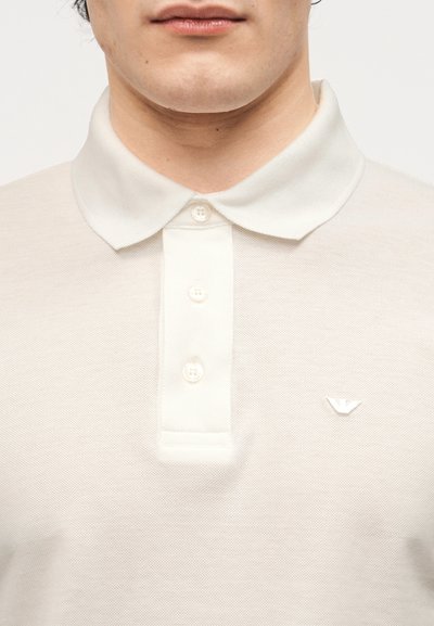 Άνδρας που φοράει λευκό πόλο μπλουζάκι με κουμπιά και μικρό κεντητό λογότυπο στο αριστερό στήθος, φαίνεται από τον λαιμό μέχρι το μέσο του κορμού.
