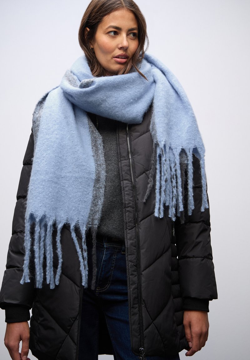 Femme portant une grande écharpe bleue claire à franges sur une veste noire matelassée, un pull gris et un jean bleu foncé sur un fond uni.