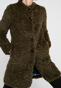 Faux-Fellmantel in Olivgrün mit einer weichen, texturierten Oberfläche. Verfügt über einen runden Kragen, fünf schwarze Knöpfe und seitliche Taschen.