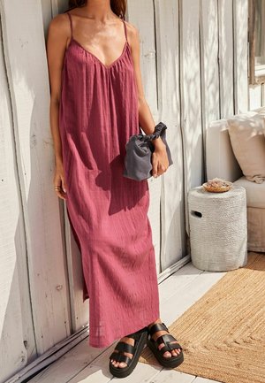 Długa, bezrękawna sukienka w kolorze różowym z głębokim dekoltem w kształcie litery V i teksturowanym materiałem. Połączona z ciemnoszarym kopertówką i masywnymi czarnymi sandałami.