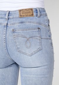 Jean en denim bleu clair avec une poche arrière ornée de coutures décoratives et une étiquette de marque en cuir marron à la taille.