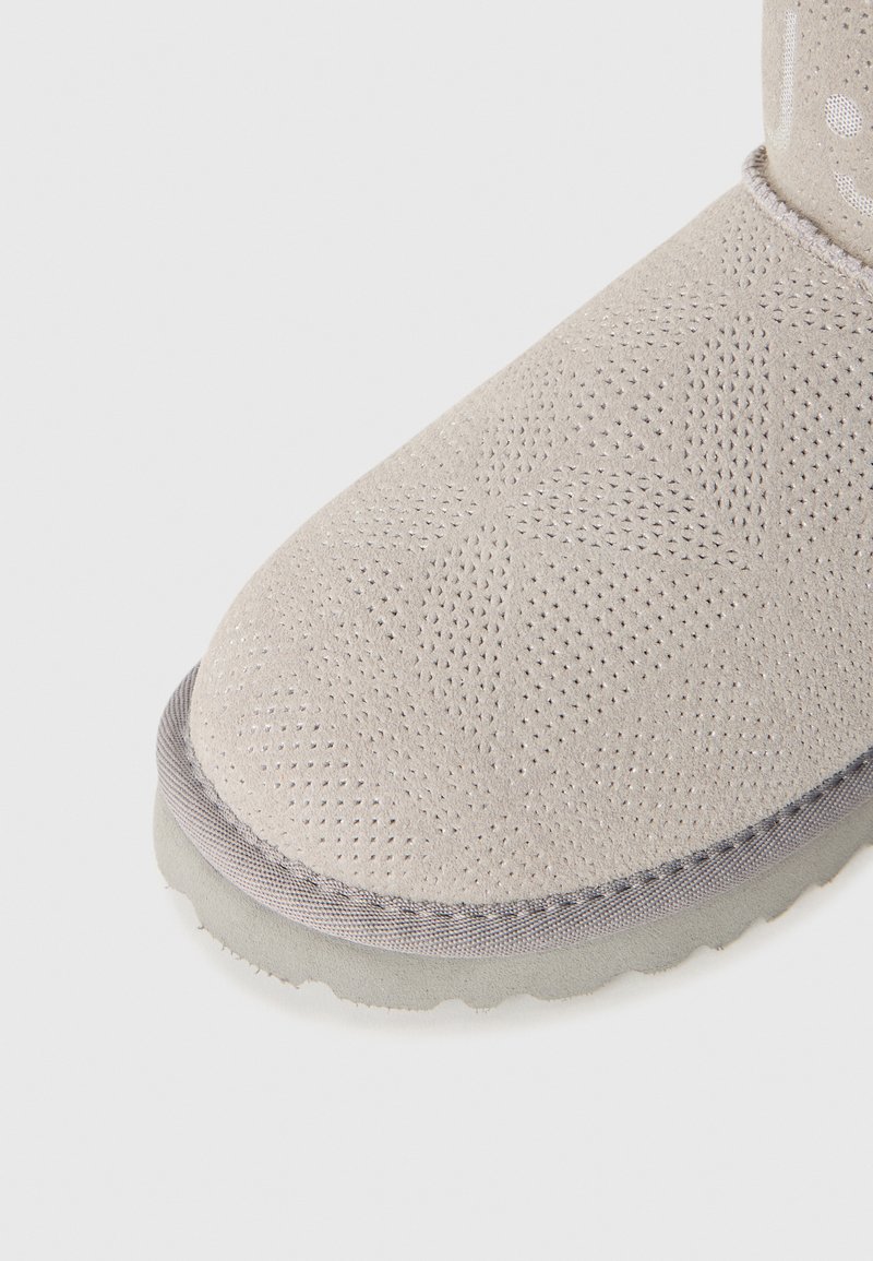 Pantoufle beige clair avec un motif géométrique perforé, une bordure grise et une semelle texturée, photographiée sur fond blanc.