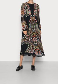 Frau in einem schwarzen Midikleid mit bunten Blumenmustern und langen transparenten Ärmeln, kombiniert mit schwarzen Loafers, stehend auf grauem Boden.