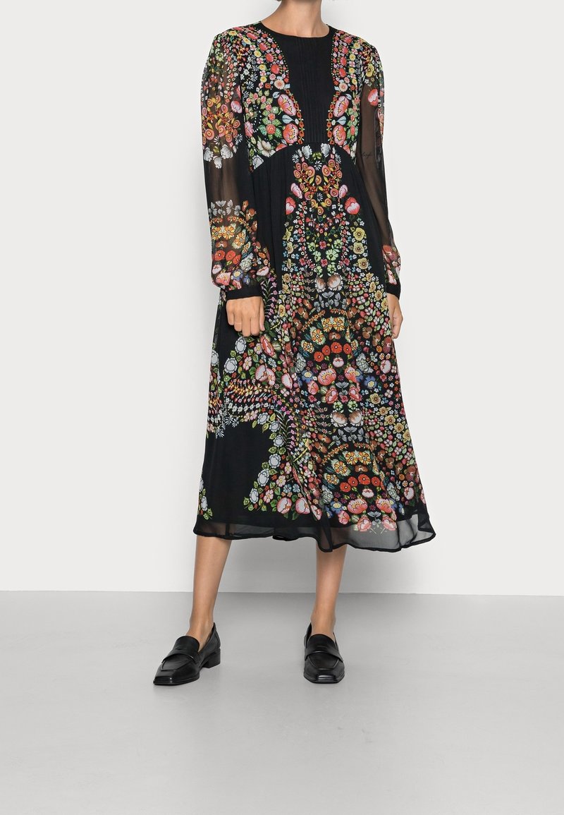 Frau in einem schwarzen Midikleid mit bunten Blumenmustern und langen transparenten Ärmeln, kombiniert mit schwarzen Loafers, stehend auf grauem Boden.