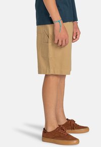 Jeune personne debout, portant un short cargo beige, des baskets marron, un tee-shirt navy et un bracelet en fil bleu au poignet gauche.