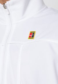 Veste blanche zippée avec un petit patch rectangulaire du logo Nike en rouge, bleu, vert et jaune sur la poitrine.