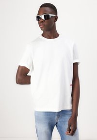Camiseta de algodón blanca con mangas enrolladas y una pequeña etiqueta roja, combinada con jeans de mezclilla azul claros y gafas de sol oscuras y elegantes.