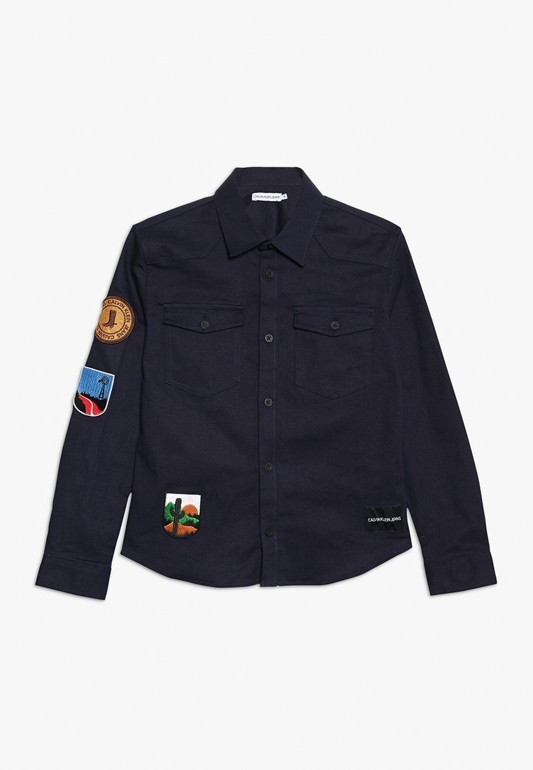 Chemise bleu marine à manches longues boutonnée avec poches poitrine et quatre patchs brodés colorés sur la manche et devant.